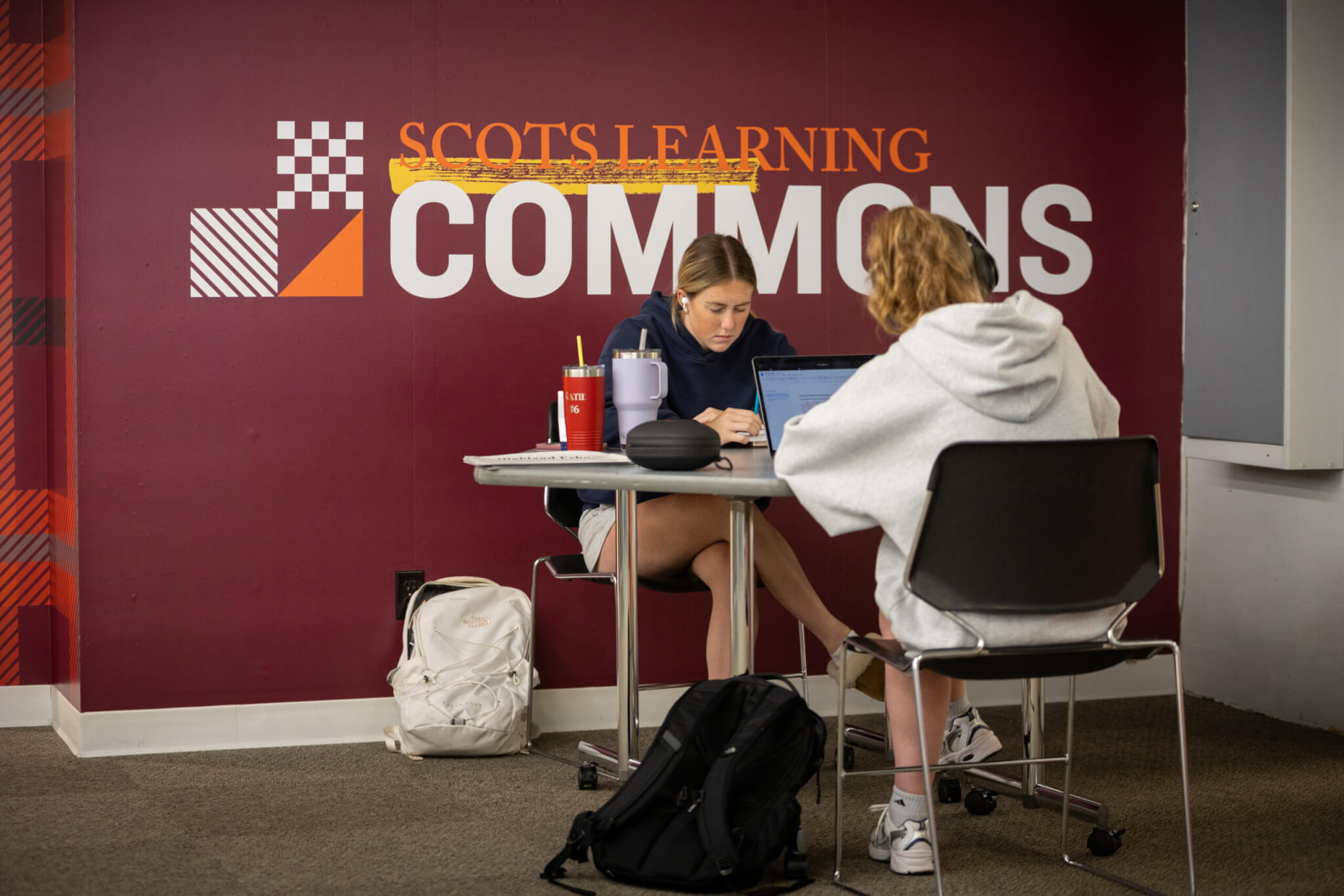 Two students study at a table in front of a Scots Learning Commons sign.