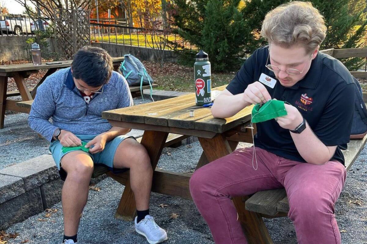 Photo of two students at a picnic table, sewing pieces of cloth.