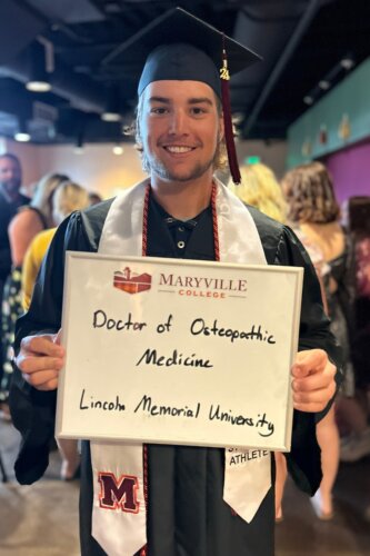 Photo of Christian Carlton holding a dry erase board in his graduation gown and mortar board