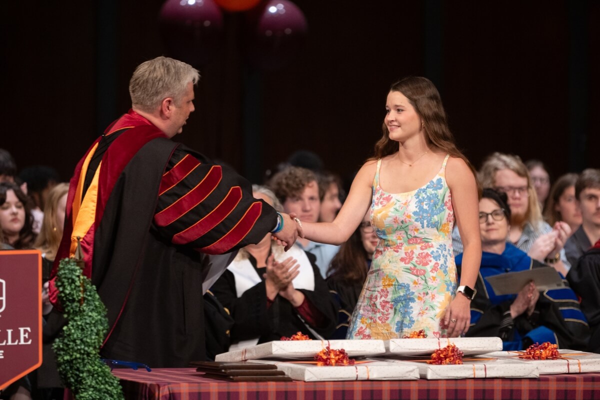 Photo of a man in academic robes congratulating a young lady by shaking her hand