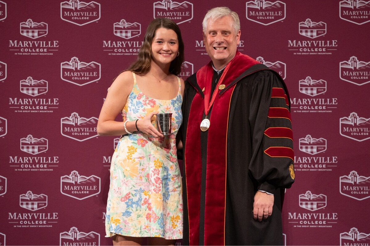 Photo of Grace McDonald holding the LeQuire Award with Dr. Bryan Coker in front of a garnet backdrop with the Maryville College logo