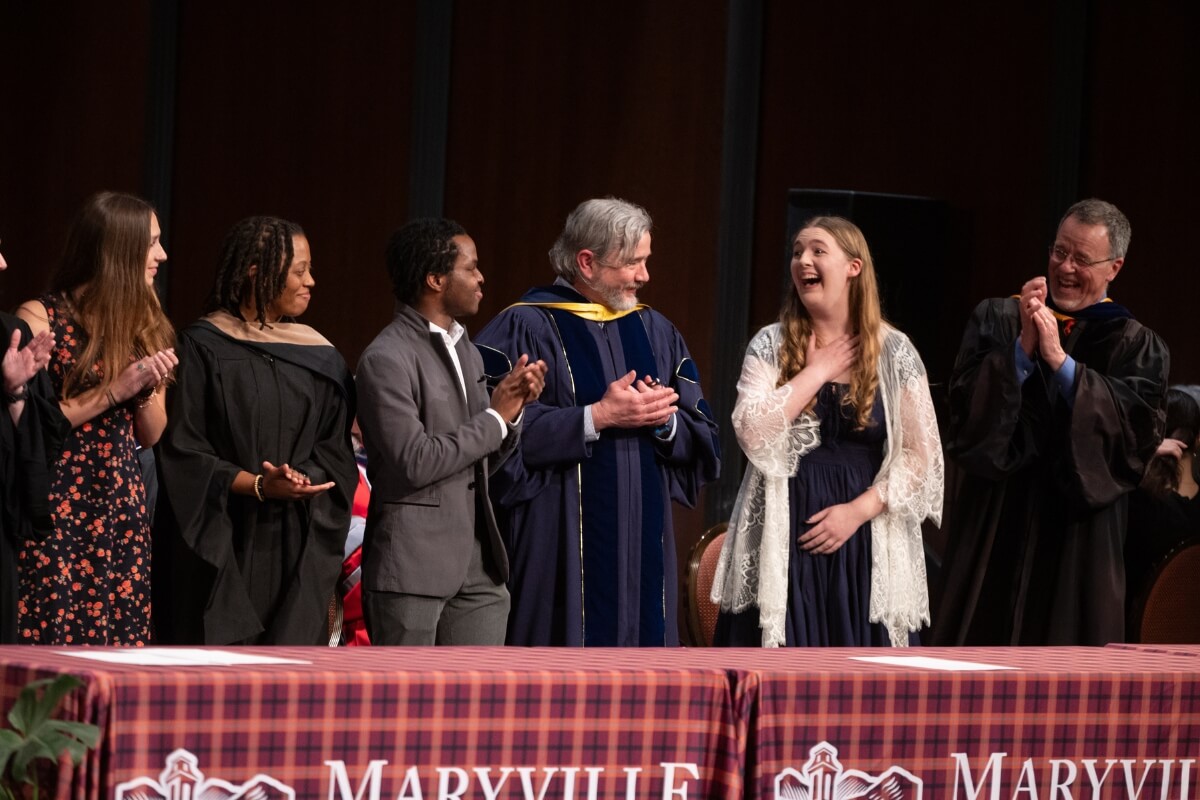 Photo of an astonished girl clutching her chest and smiling surrounded by peers