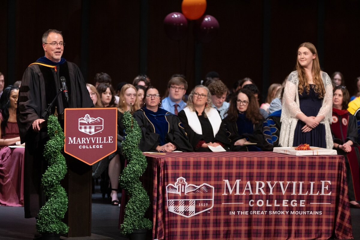 Photo of a professor in academic robes at a podium speaking while a girl to his left smiles and listens