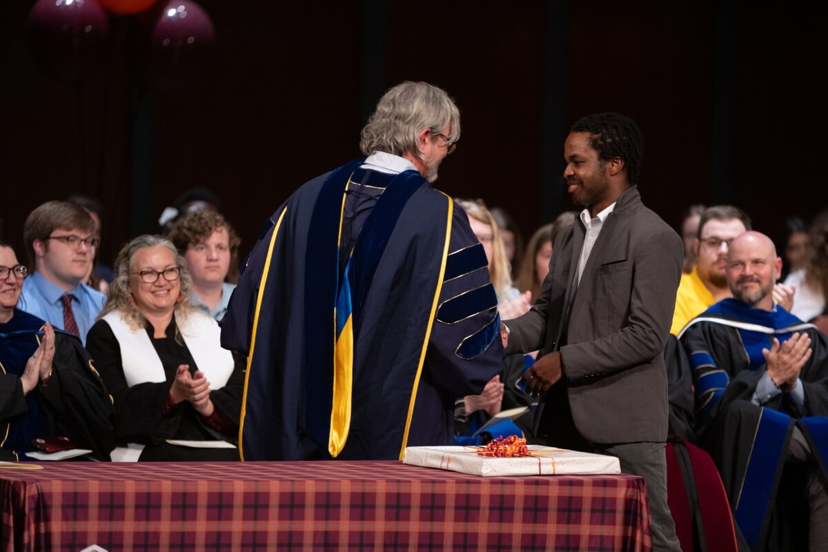 Photo of a professor in an academic gown handing a Black man a package