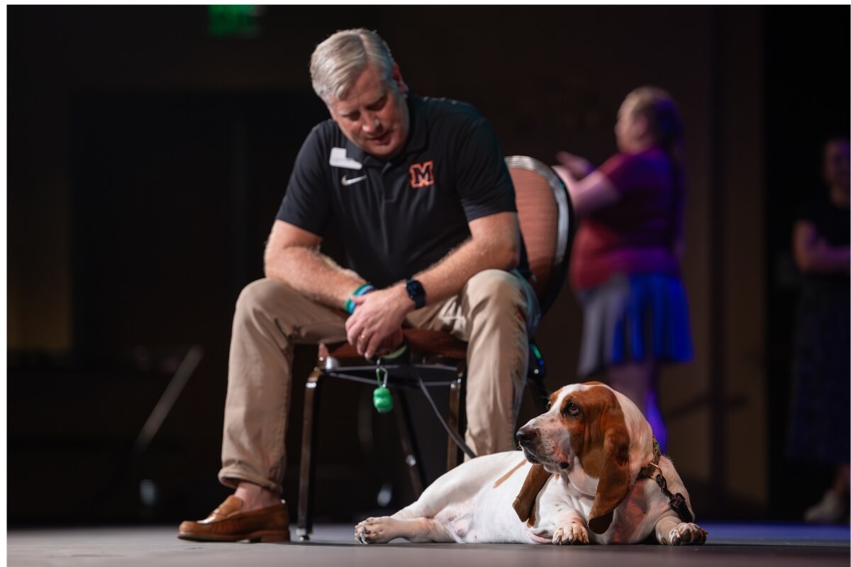Photo of President Bryan Coker seated on a stage with his Basset hound, Dolly, at his feet