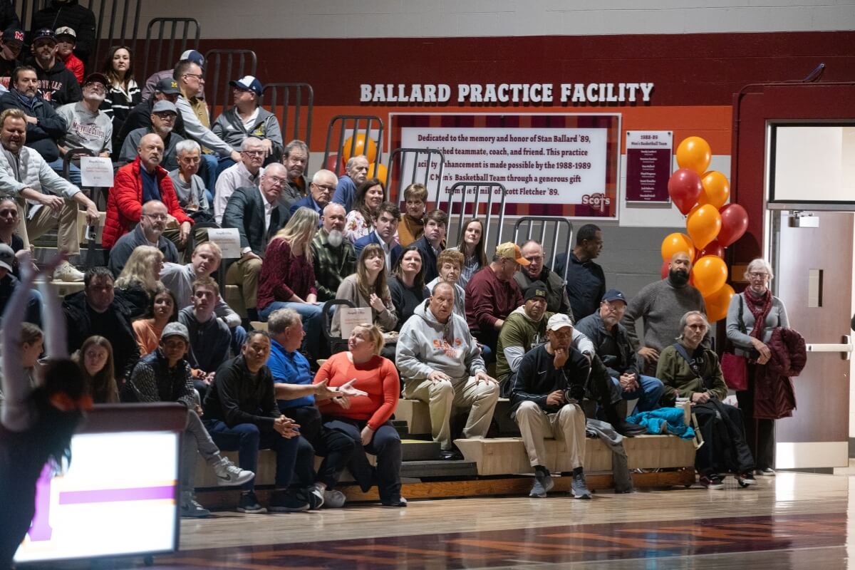 Photo of a crowd sitting in bleachers beneath a Ballard Practice Facility sign