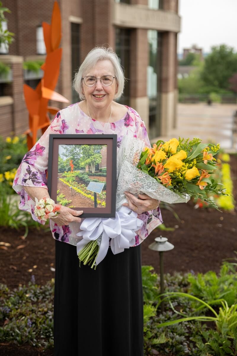 Photo of Sally Gross holding a painting and flowers in front of the Clayton Center memorial garden