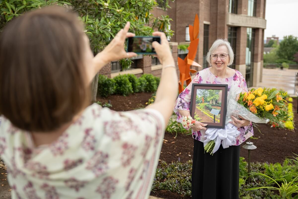 Photo of someone taking Sally Gross' picture in front of the Clayton Center memorial garden