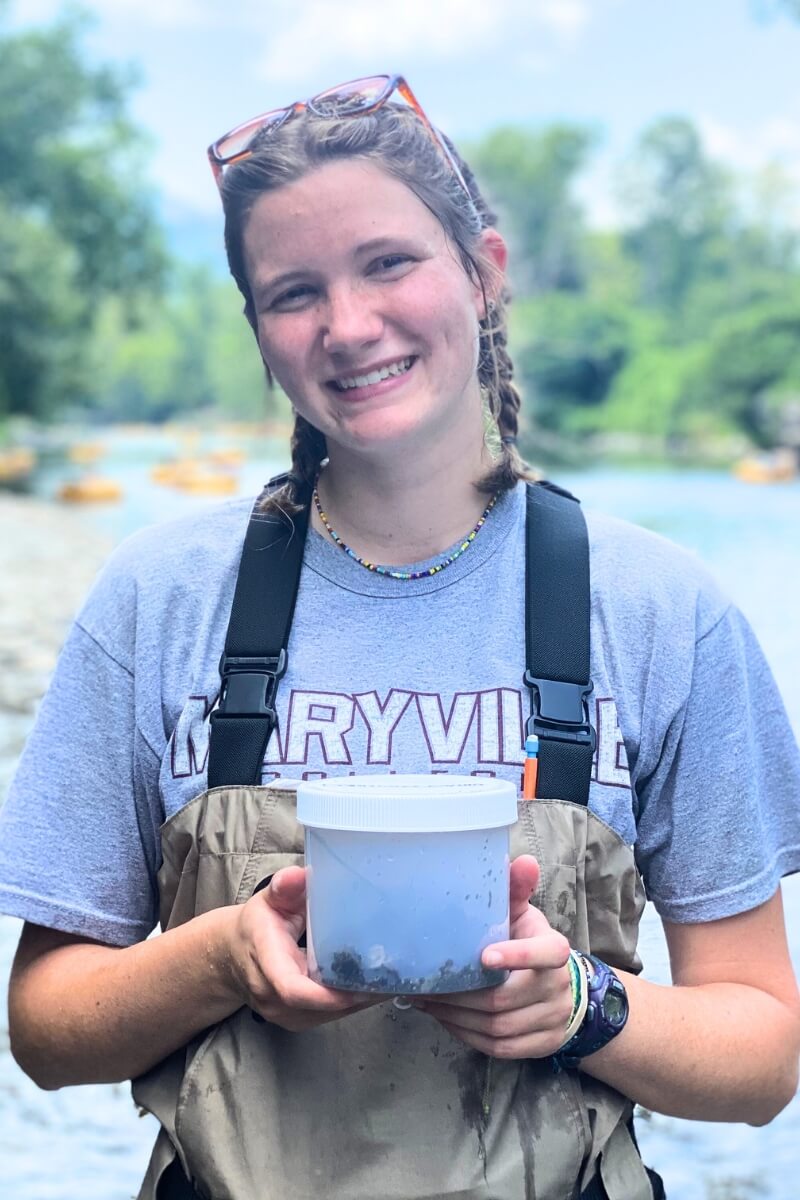 Photo of recent Maryville College graduate Kaitlin Koster '25 smiling while she holds a sample of macroinvertebrates from the Little River.