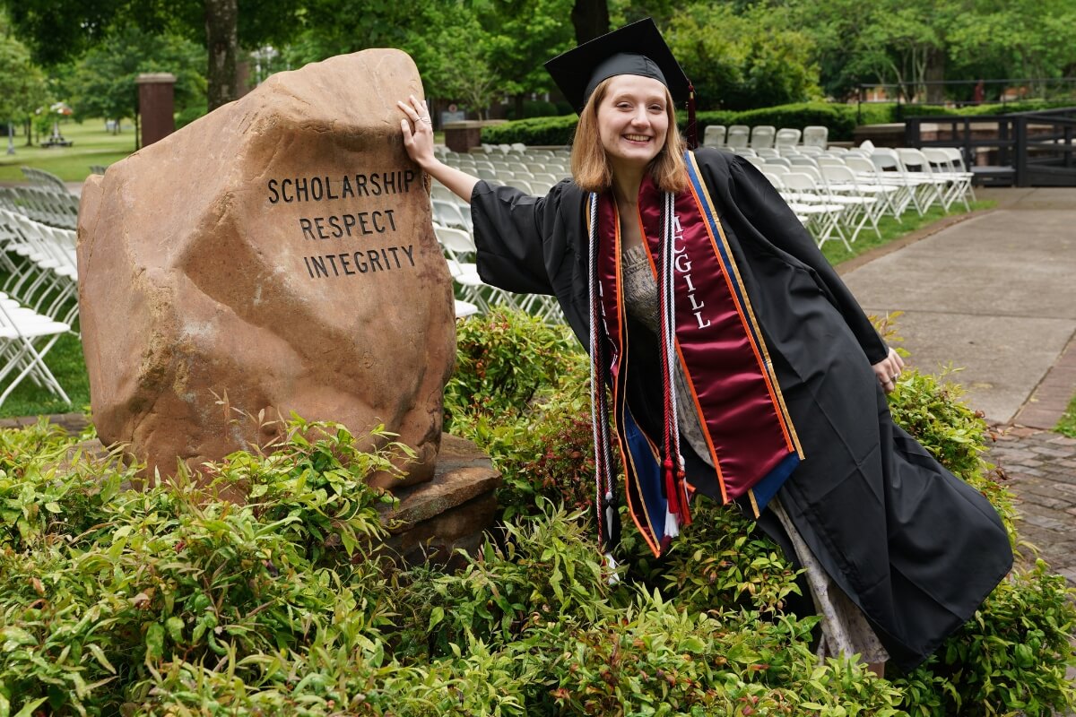 Photo of a smiling Maryville College graduate in cap and gown touching the Covenant Stone