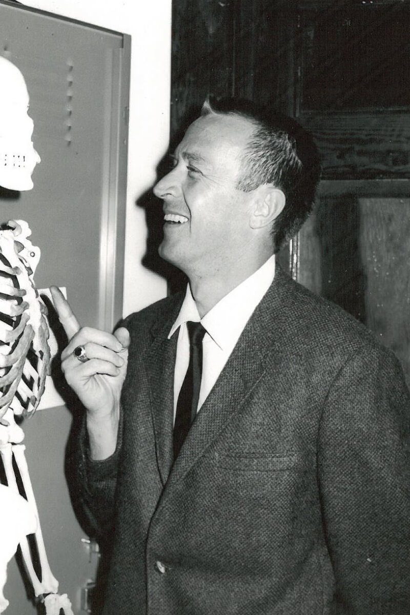 Black and white photo of a man in a suit pointing to a skeleton with a grin on his face