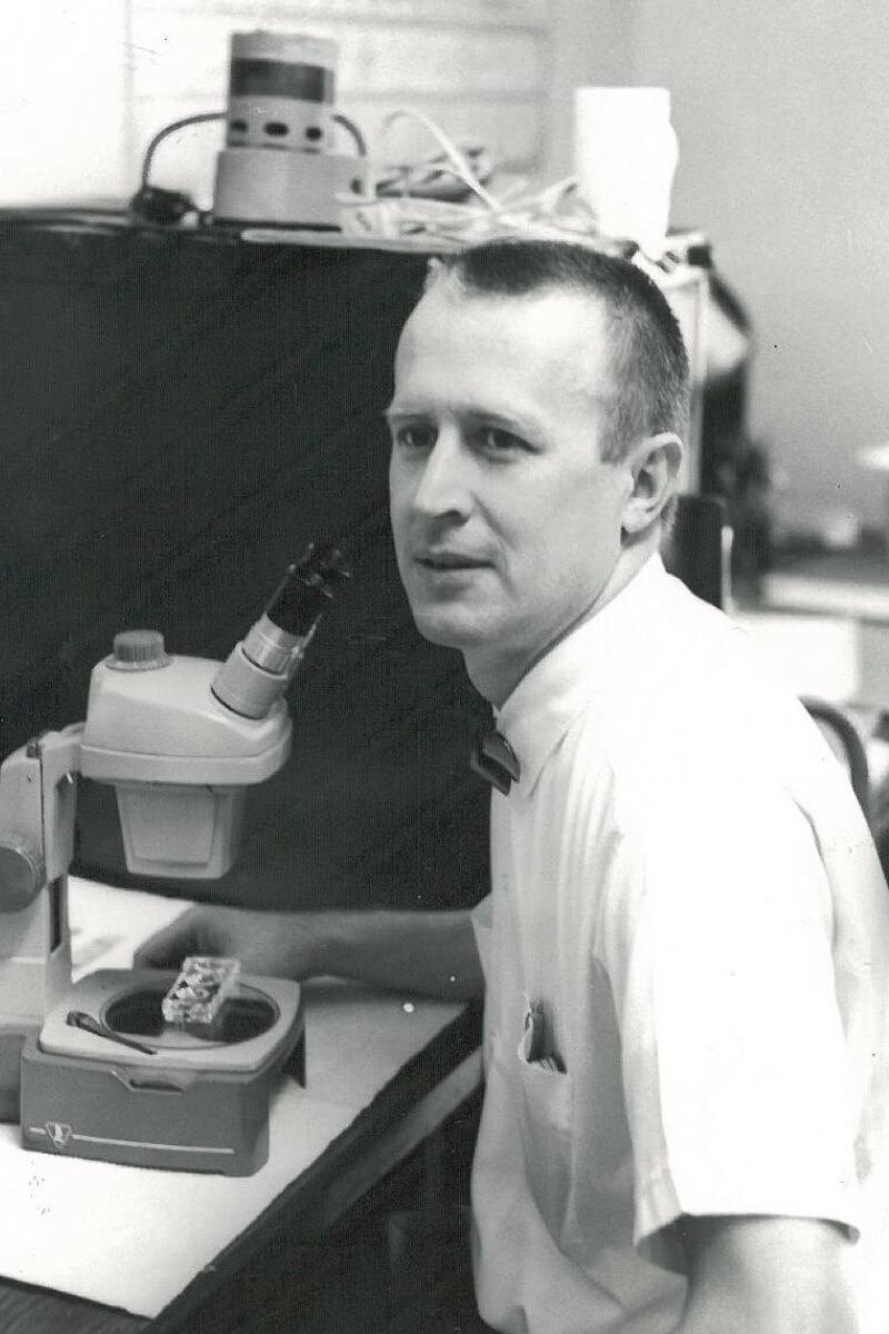 Black and white photo of a scientist sitting at a microscope turning to look at the camera