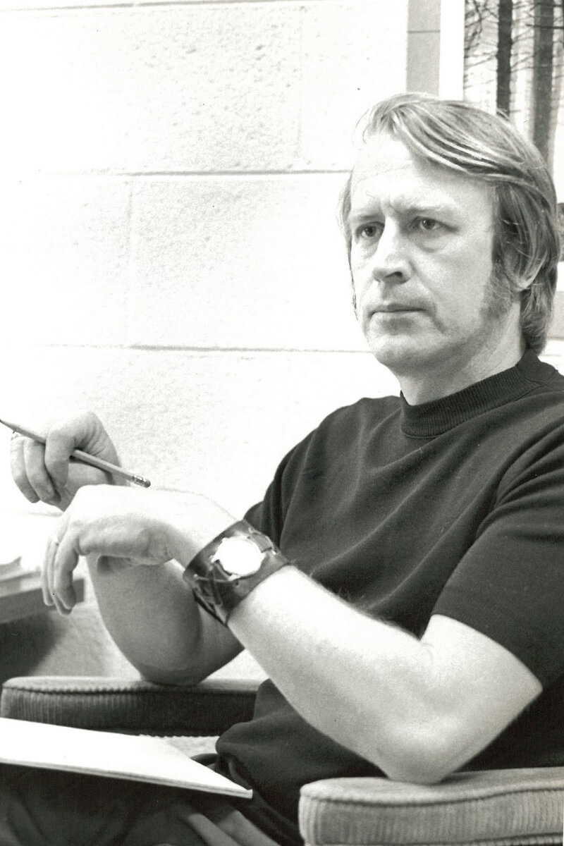 Photo of a mustachioed man sitting in a desk chair, clutching a pencil