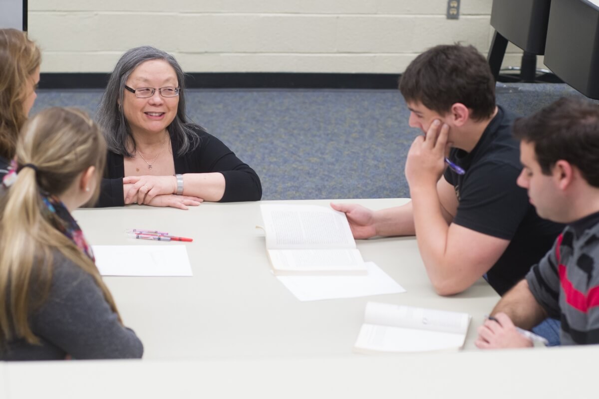 Photo of Dr. Kathie Shiba, kneeling at a table full of students and smiling