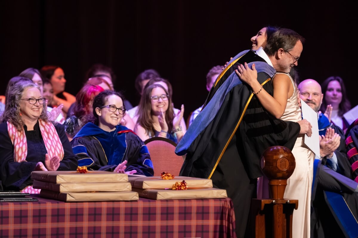 Photo of a student embracing a faculty member in graduation regalia