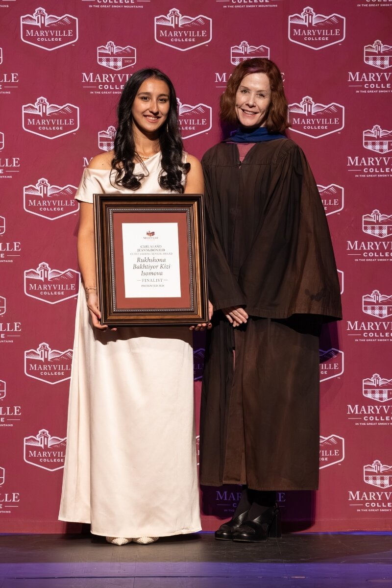 Photo of a smiling girl holding a plaque with a professor beside her in regalia