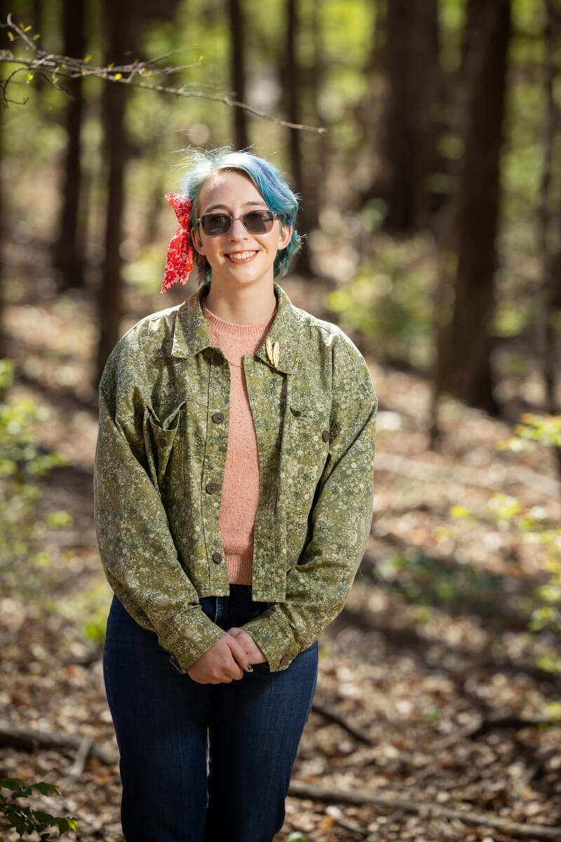 Photo of Gianna Mantegna smiling and standing in dappled sunlight in the Maryville College Woods.