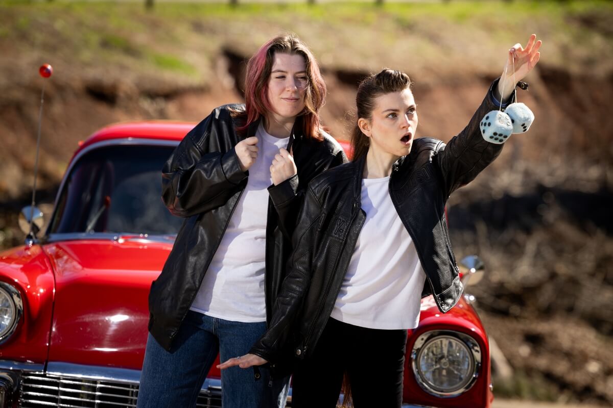 Photo of two students in front of a classic car, one holding up a pair of fuzzy dice