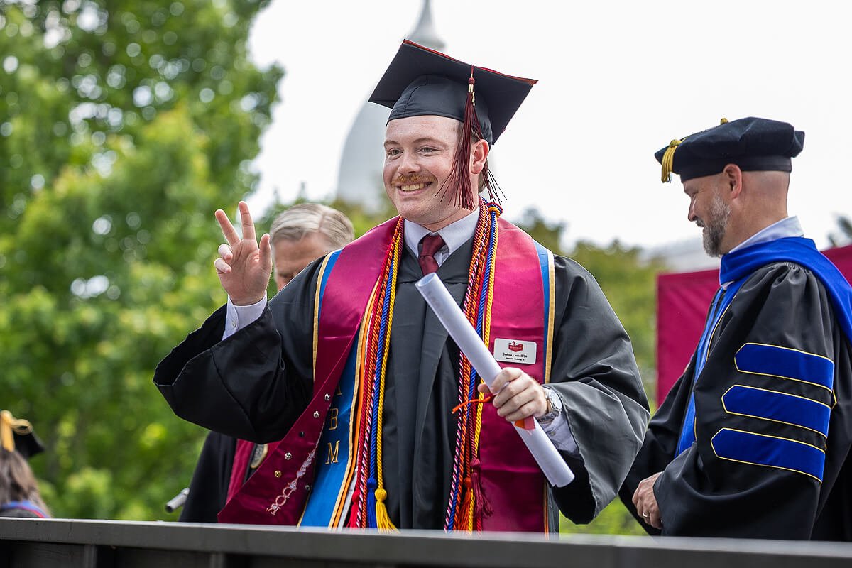 Photo of a young man smiling and flashing a peace sign while holding a diploma