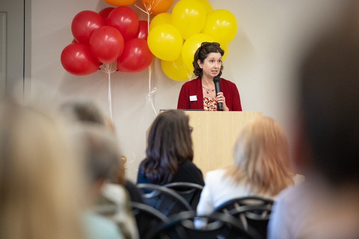 Photo of a woman wearing a red jacket standing at a podium with balloons behind her