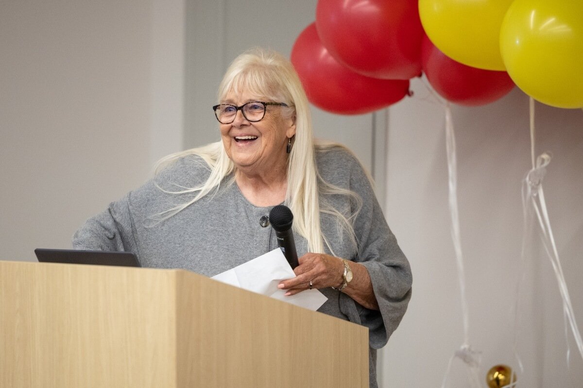 Photo of a woman with blonde hair talking into a microphone at a podium beside a bunch of balloons