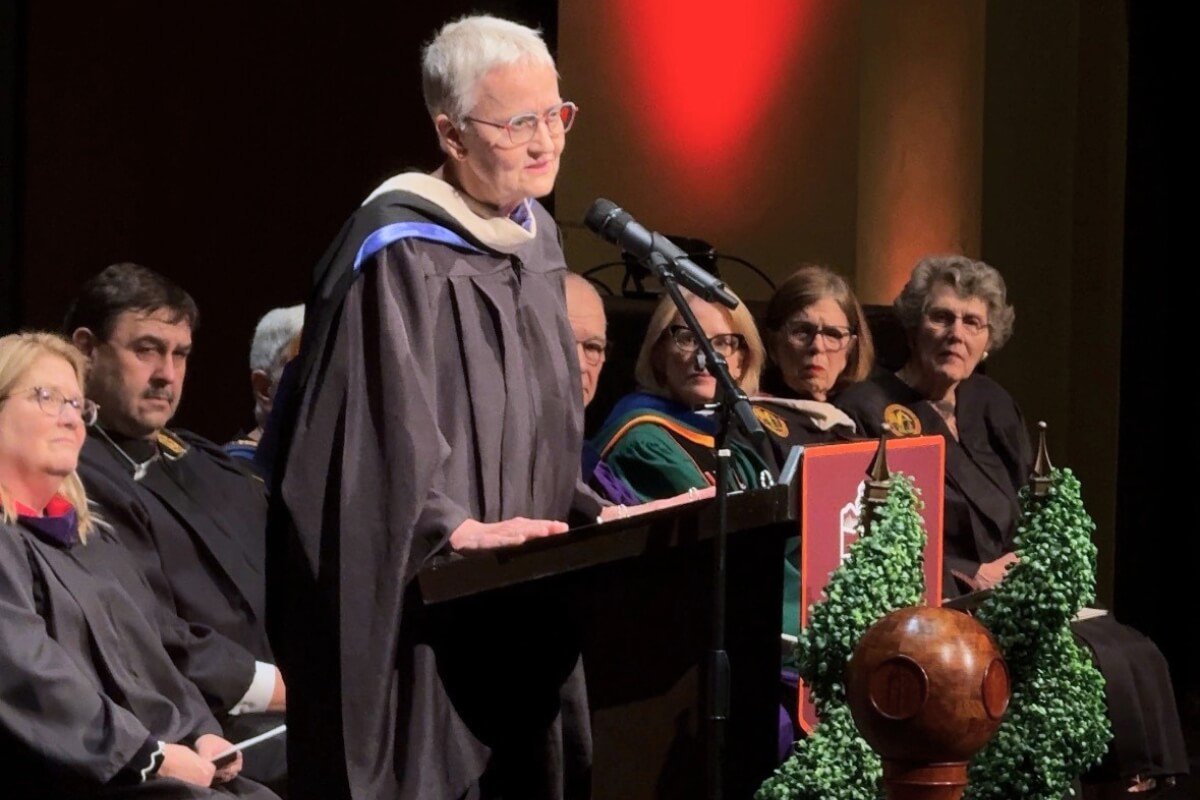 An elderly woman wearing graduation regalia speaks at a podium